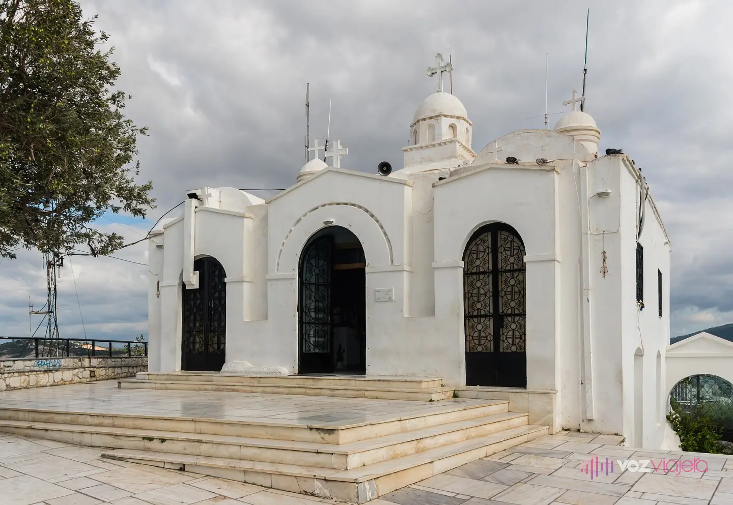 La capilla encalada de Ágios Geórgios en la cima del Licabeto, con sus cúpulas blancas, el campanario, las puertas de reja abiertas y el atrio de mármol de la entrada