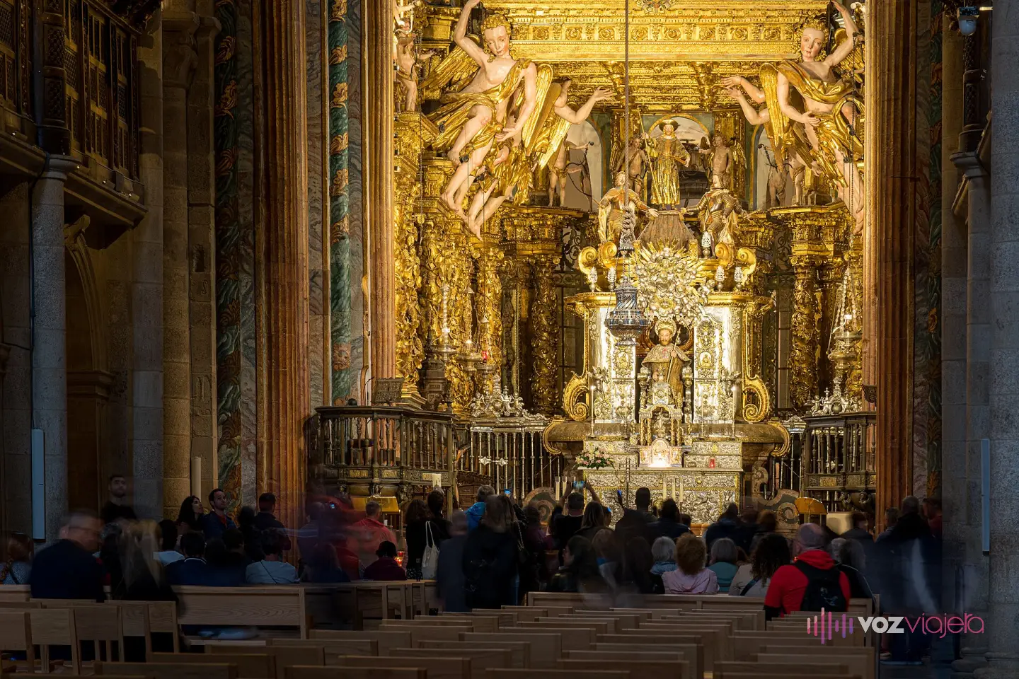 El altar mayor barroco de la catedral de Santiago, dorado y con ángeles sobre el baldaquino, con visitantes sentados en los bancos de la nave