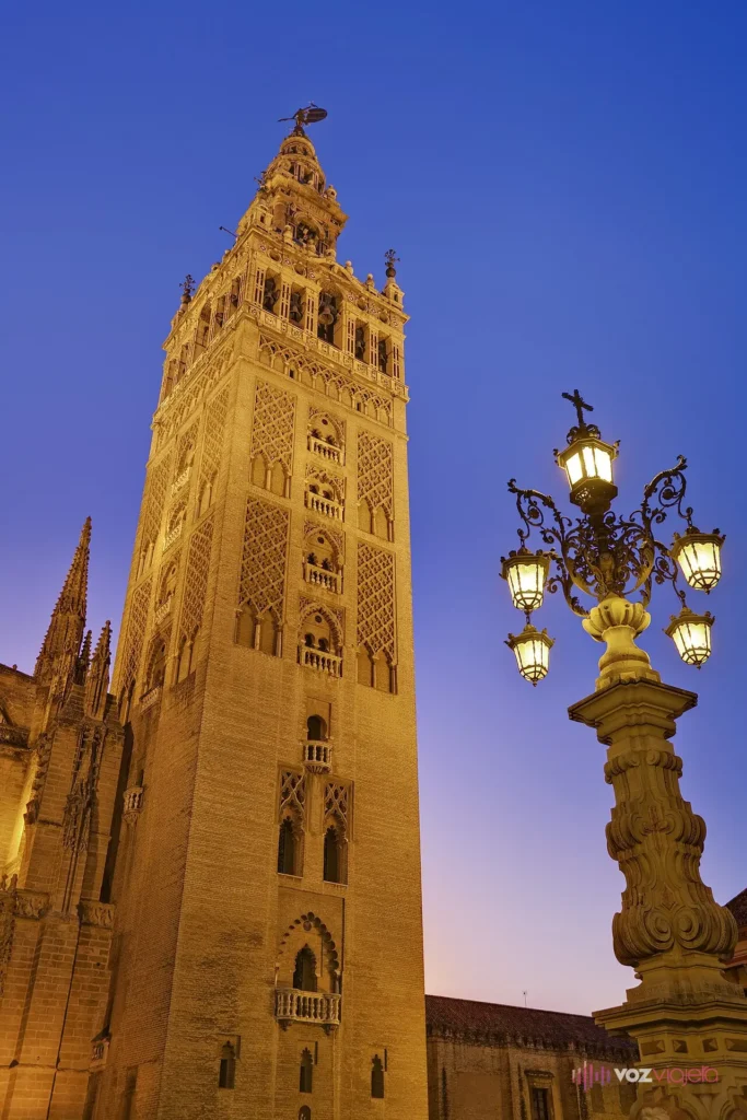 La Giralda de la Catedral de Sevilla iluminada al anochecer sobre un cielo azul intenso, con una farola de hierro forjado en primer plano