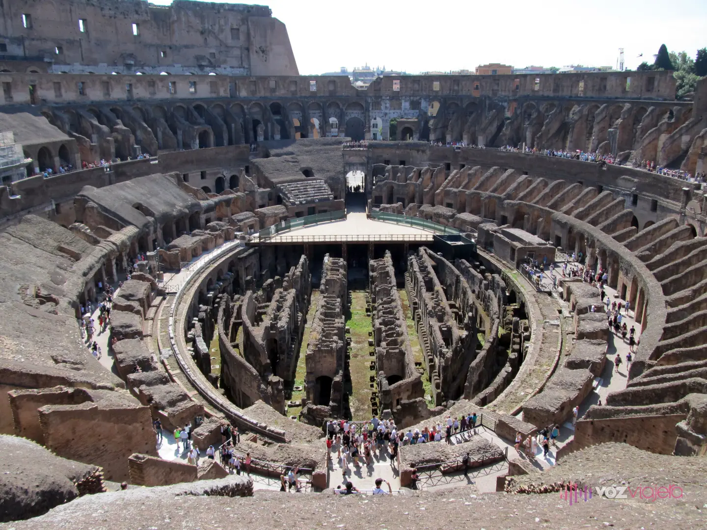 Interior del Coliseo visto desde lo alto de las gradas: en el centro, el hipogeo al descubierto, un laberinto ovalado de muros y pasillos de ladrillo bajo el nivel de la arena, con una plataforma clara que cubre uno de sus extremos y visitantes caminando por el borde; alrededor, los restos escalonados de la cávea