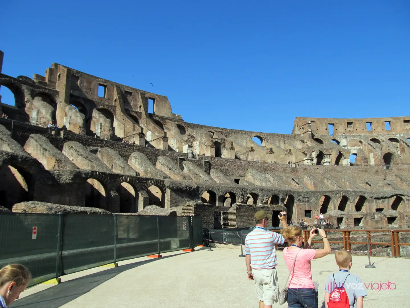Visitantes caminando sobre la plataforma que cubre parte de la arena del Coliseo, una familia de espaldas hace fotos con el móvil; al fondo, las gradas en ruinas y los arcos de los pisos superiores bajo un cielo azul
