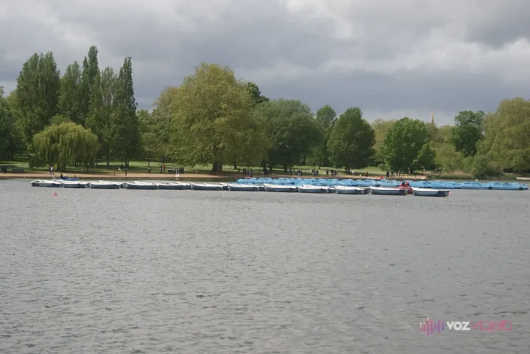 El lago Serpentine de Hyde Park con una fila de barcas de alquiler azules amarradas en la orilla de enfrente, bajo un cielo nublado y con árboles altos detrás