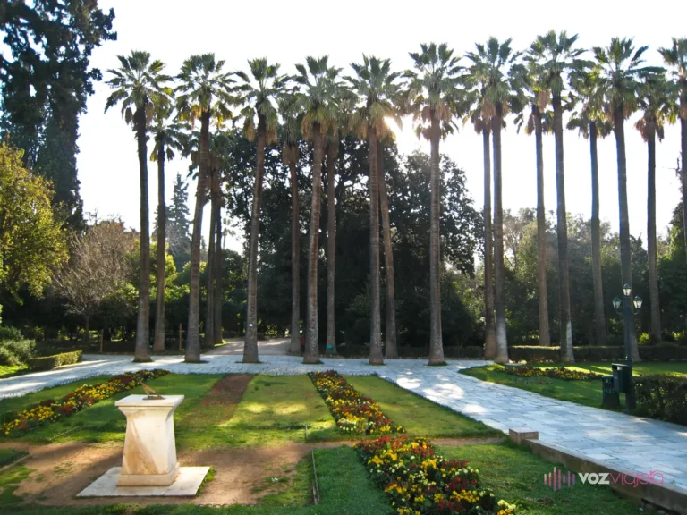 Avenida de palmeras altísimas del Jardín Nacional de Atenas a contraluz, con un camino de losas de mármol, parterres de flores amarillas y rojas y, en primer plano, un reloj de sol sobre un pedestal de mármol blanco