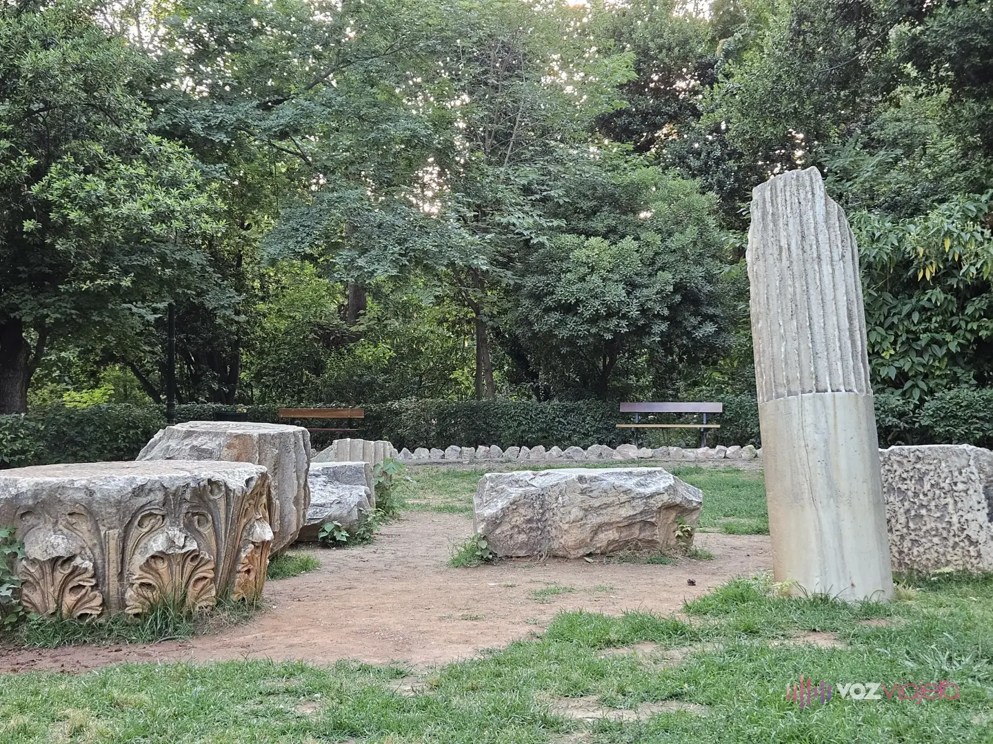 Restos antiguos de mármol sobre el césped del Jardín Nacional de Atenas: a la izquierda, un capitel corintio con hojas de acanto talladas; a la derecha, un fuste de columna acanalada en pie, con bancos y árboles al fondo