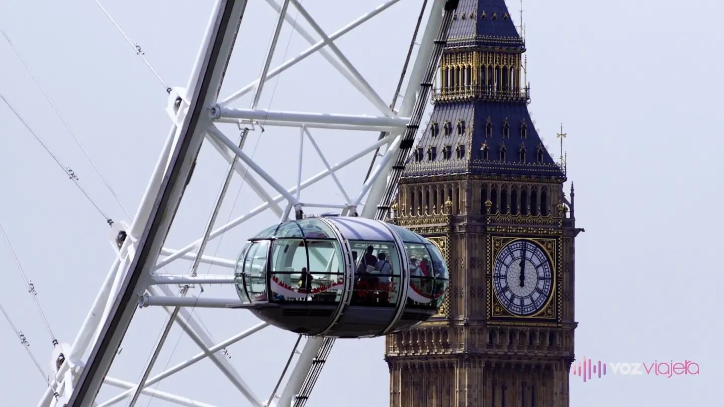 Una cápsula ovalada de cristal del London Eye con visitantes dentro, colgada del aro blanco de la noria, y detrás la torre del Big Ben con la esfera del reloj