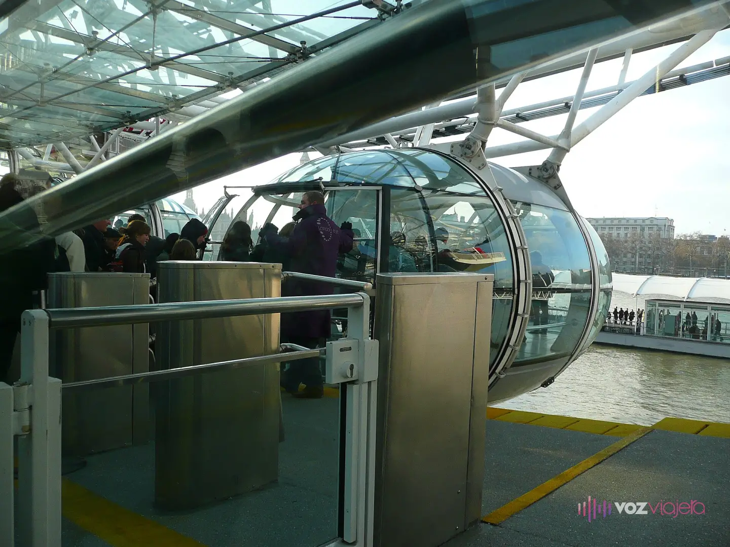 Andén de embarque del London Eye bajo una cubierta de cristal: visitantes entrando en una cápsula a través de unos tornos metálicos, con un empleado junto a la puerta y el Támesis al fondo