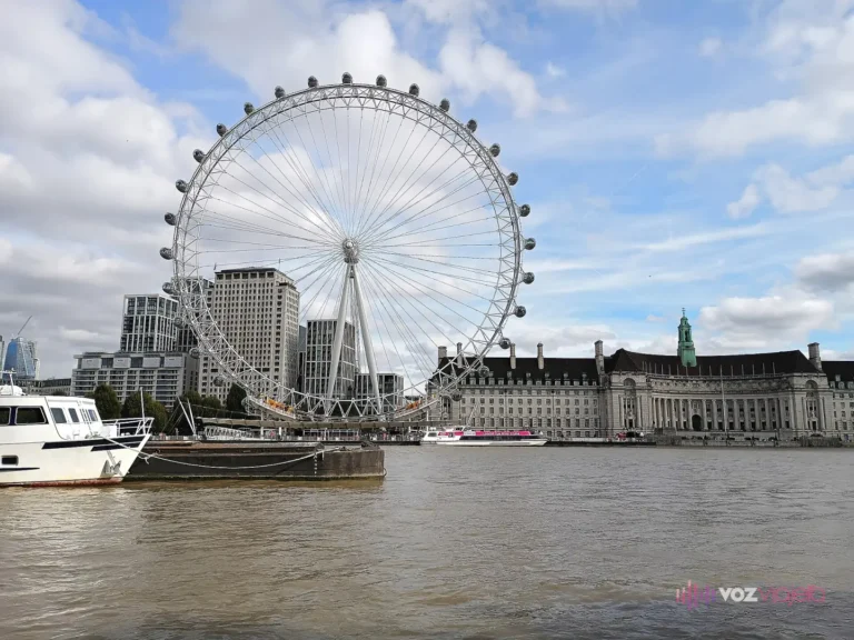 El London Eye entero en la orilla sur del Támesis en un día de nubes y claros: la rueda blanca con sus cápsulas, el edificio de County Hall a la derecha y un barco amarrado en primer plano