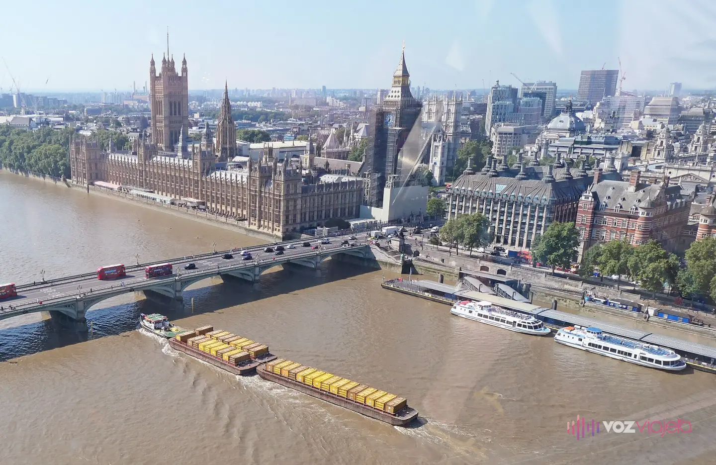 Vista desde una cápsula del London Eye del Palacio de Westminster y la torre del Big Ben, con andamios, sobre el Támesis, el puente de Westminster con autobuses rojos y una barcaza de contenedores amarillos