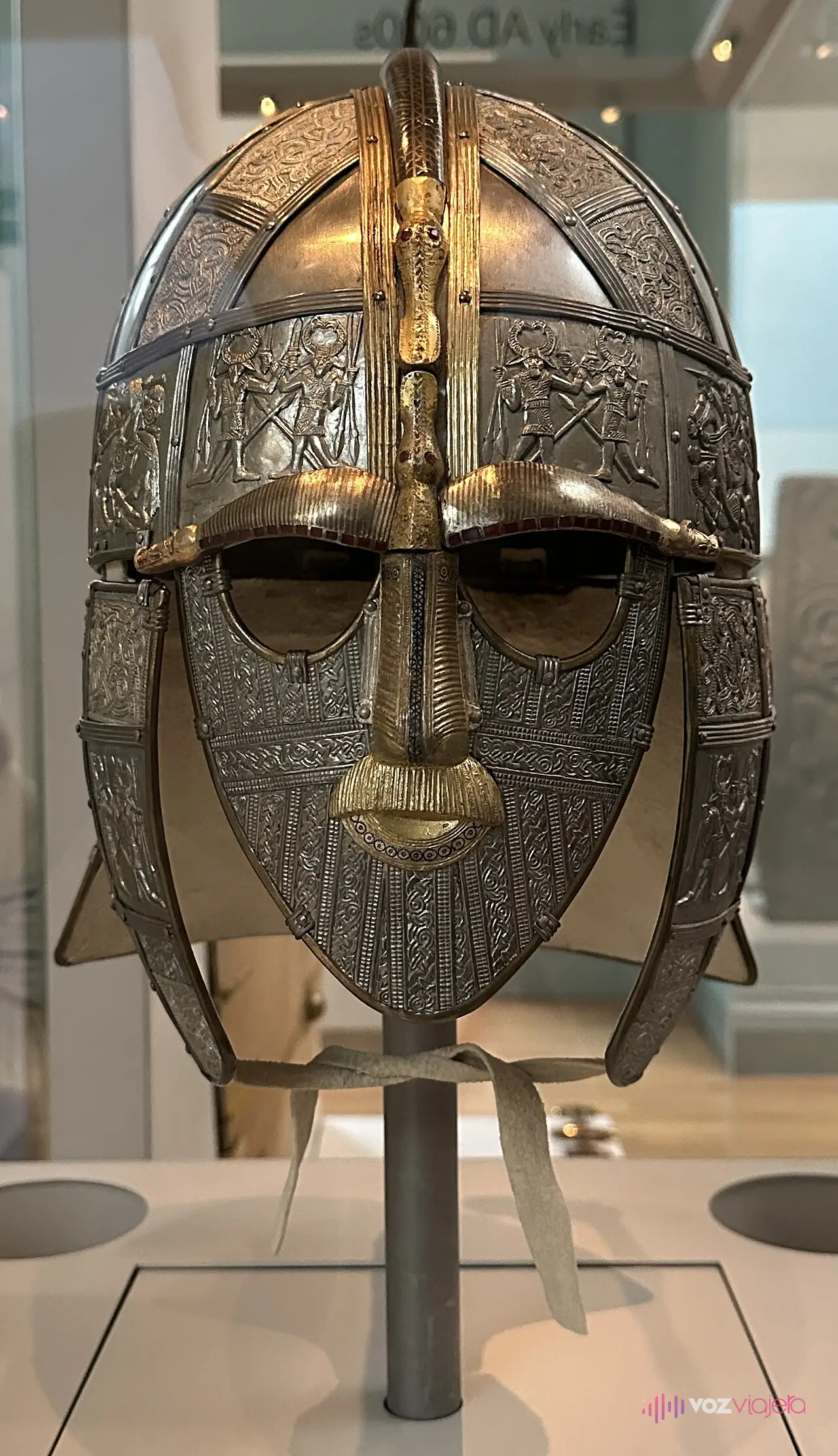 Réplica del casco de Sutton Hoo en su vitrina de la sala 41 del Museo Británico: casco de hierro con cara completa, cejas y nariz doradas, bigote y placas decoradas con guerreros
