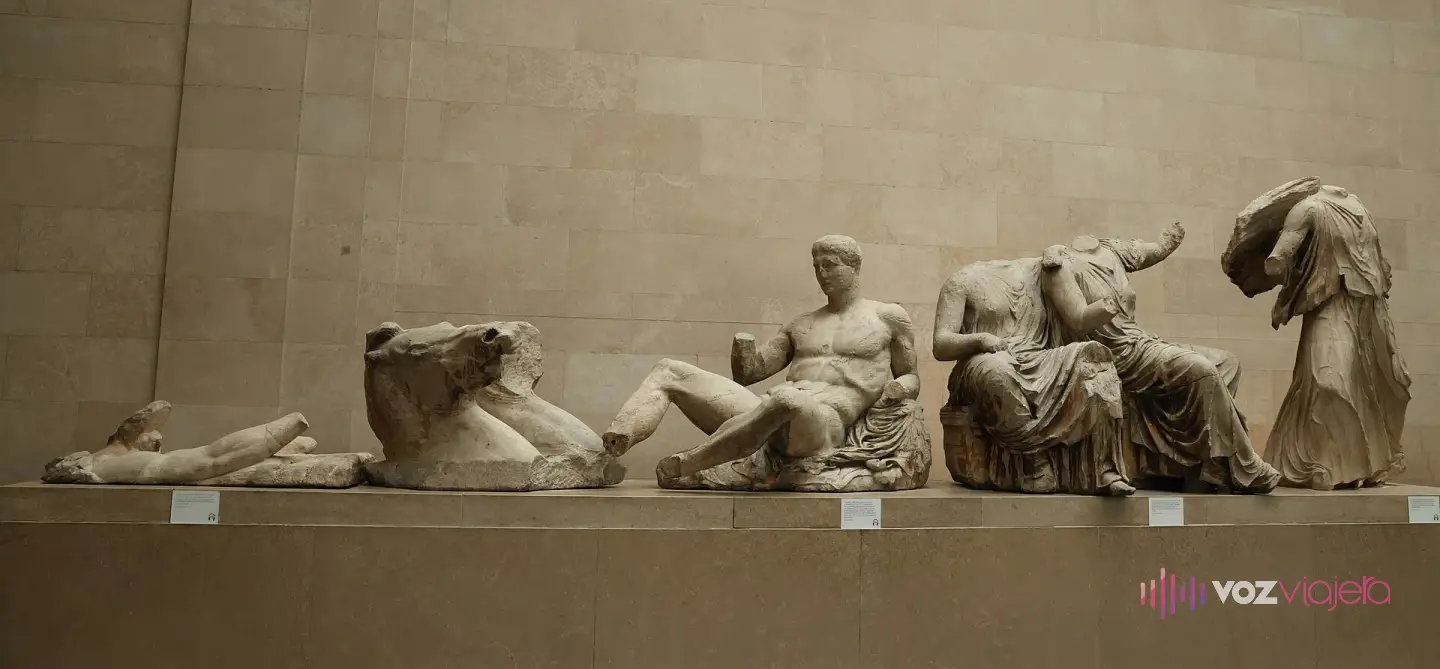 Figuras de mármol de un frontón del Partenón expuestas en fila sobre un pedestal en la sala 18 del Museo Británico: una cabeza de caballo, un joven recostado y 3 figuras femeninas sin cabeza