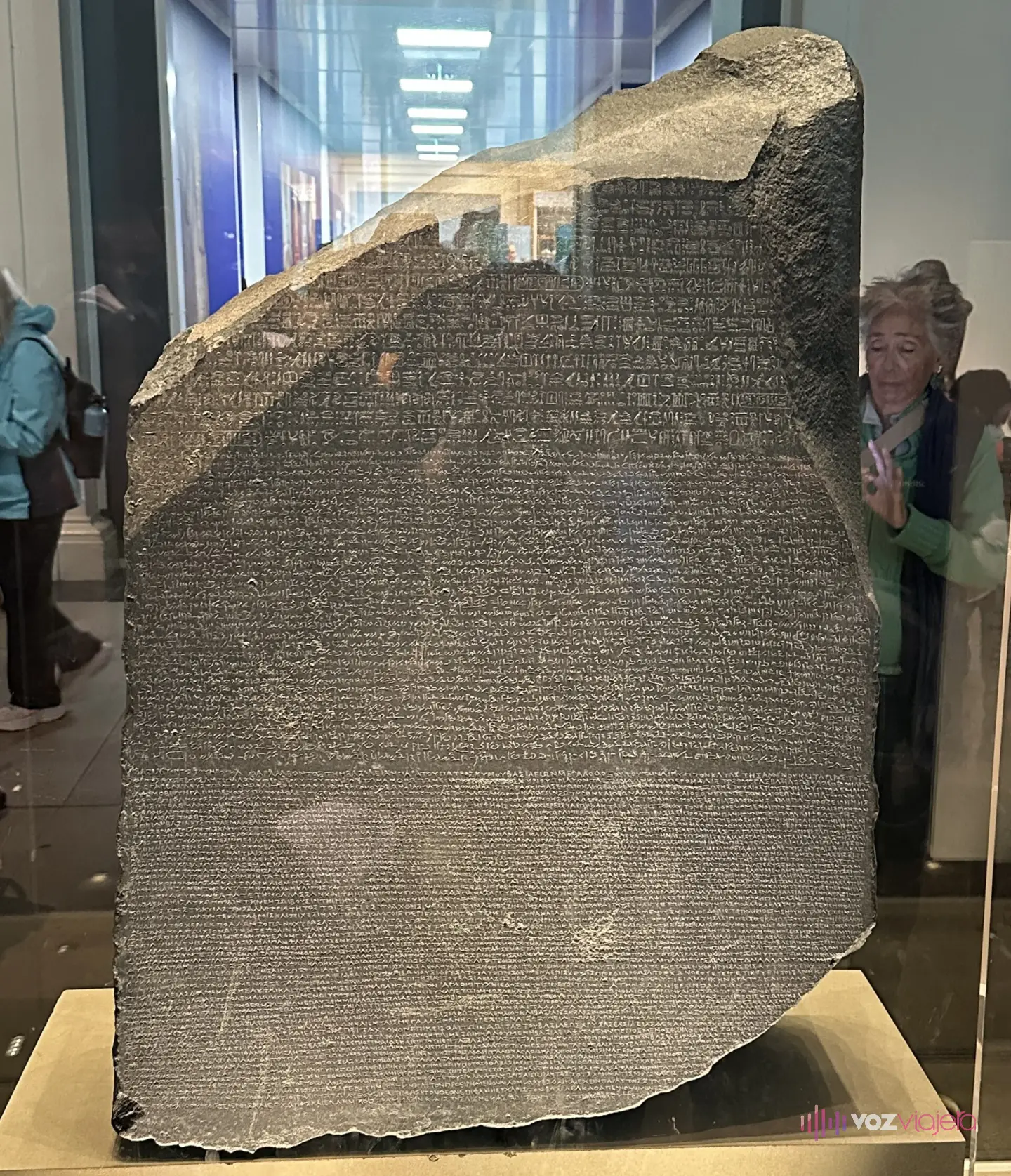 La Piedra de Rosetta, un bloque de piedra oscura con la esquina superior rota y el frente cubierto de líneas de escritura grabada, en su vitrina de la sala 4 del Museo Británico con visitantes detrás