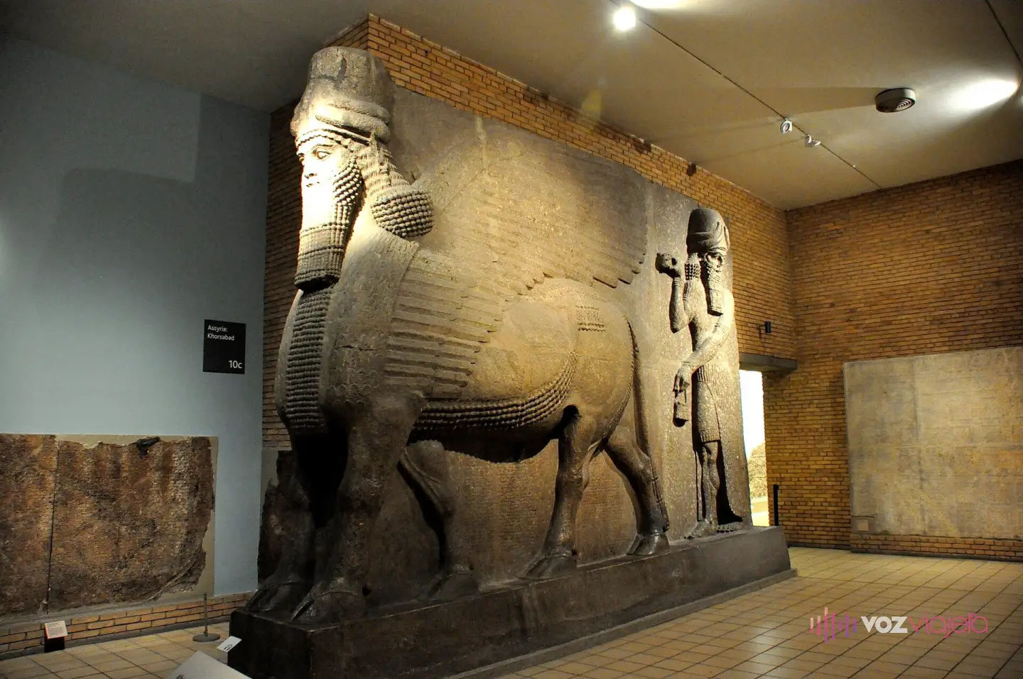 Toro alado con cabeza humana y barba rizada del palacio de Sargón II en Jorsabad, tallado en un gran bloque de piedra junto a una figura divina alada, en la sala 10 del Museo Británico