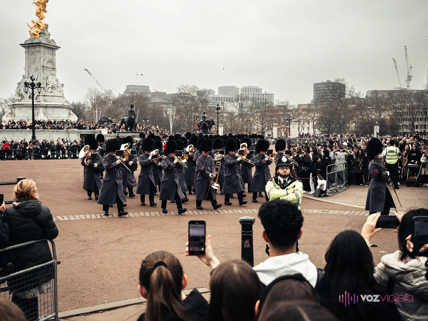 Banda de la Guardia con gorros de piel de oso y capotes grises desfilando junto al Monumento a Victoria durante el cambio de guardia, con cientos de personas detrás de las vallas grabando con el móvil y un policía en primer plano