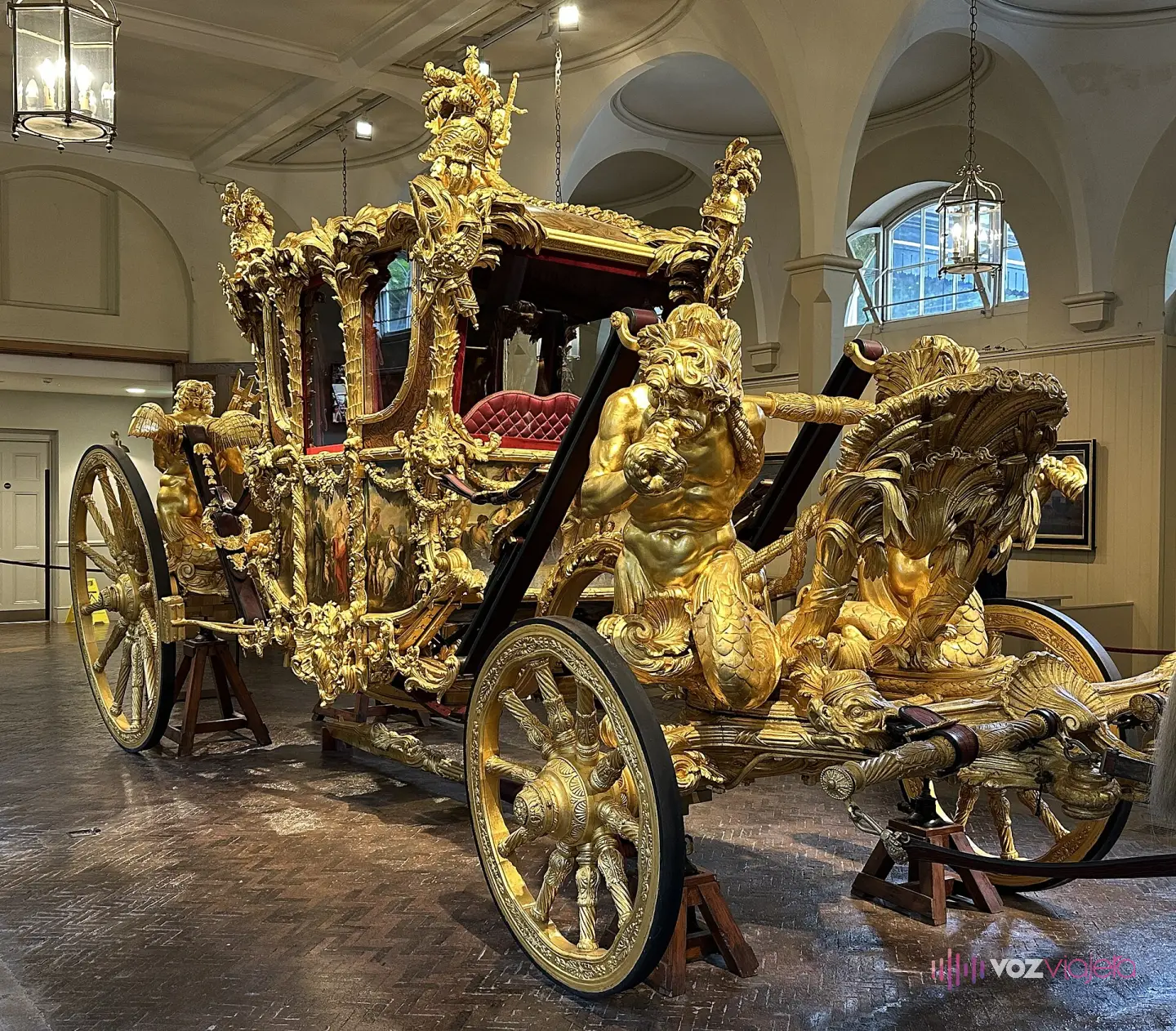 La Carroza de Oro expuesta en una sala abovedada de las Caballerizas Reales: carruaje dorado con tritones tallados en la parte delantera, paneles pintados en los costados, asiento de terciopelo rojo y 4 ruedas doradas sobre caballetes