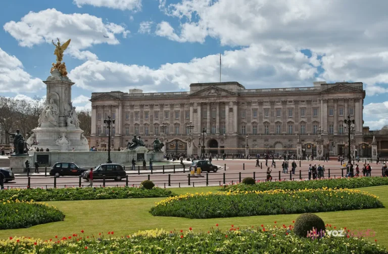 La fachada este del palacio de Buckingham con el balcón en el cuerpo central y la bandera en lo alto, las verjas negras y doradas, el Monumento a la Reina Victoria con su figura dorada a la izquierda y parterres de tulipanes en primer plano