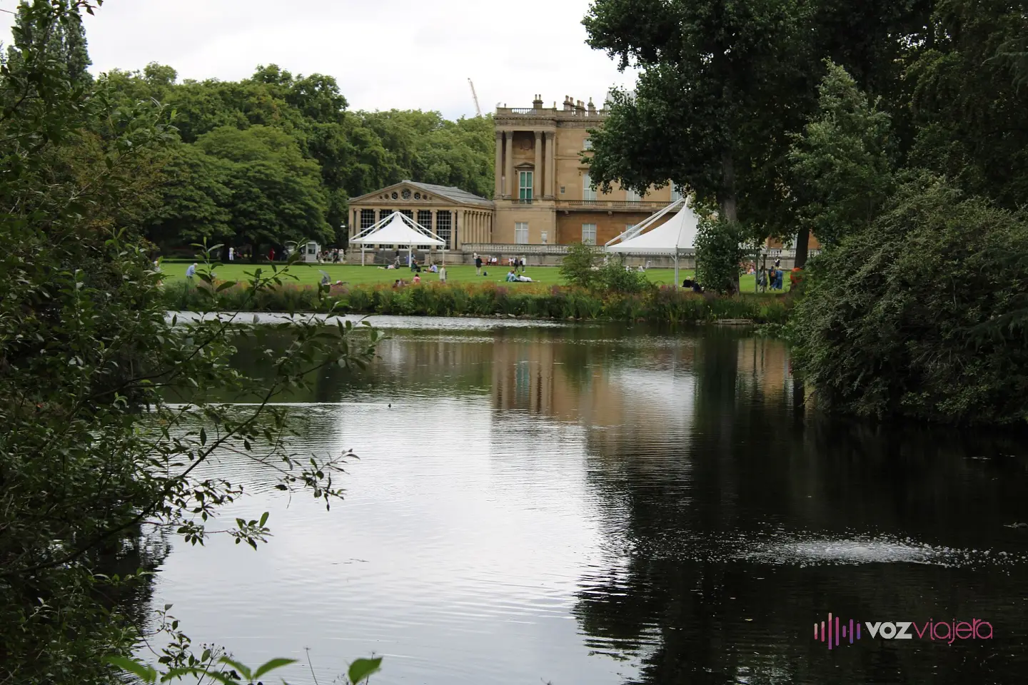 El lado del palacio de Buckingham que da al jardín, visto desde la otra orilla del lago, con el césped, 2 carpas blancas y visitantes paseando y sentados en la hierba