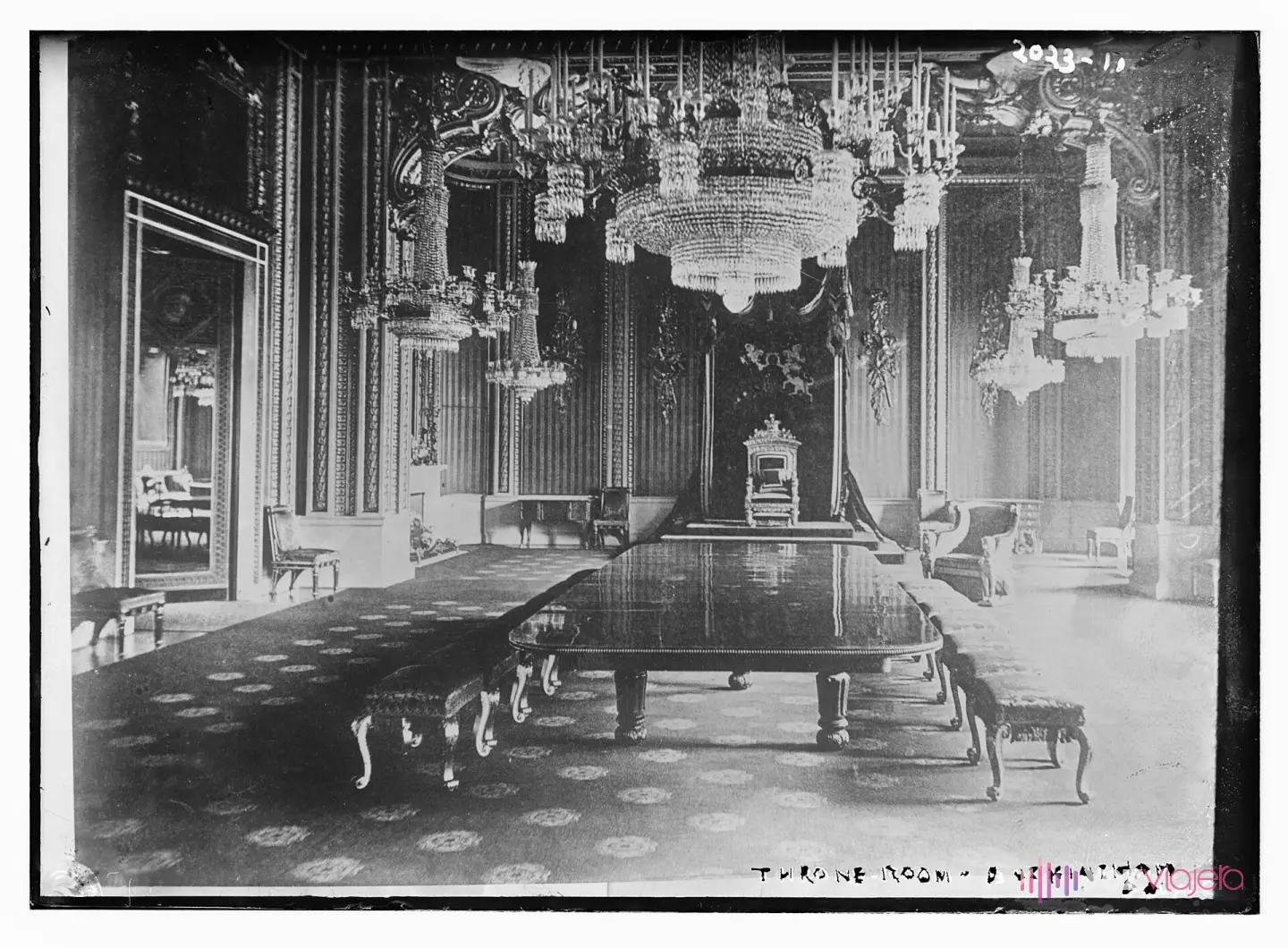 Fotografía antigua en blanco y negro del Salón del Trono del palacio de Buckingham: el trono sobre una tarima bajo un dosel con el escudo real, grandes lámparas de araña de cristal y una mesa larga con bancos en primer plano