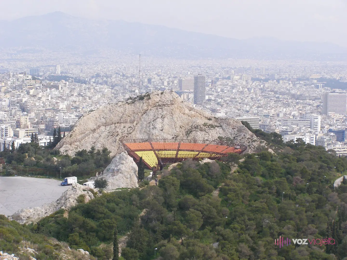 El teatro al aire libre del Licabeto visto desde arriba, con sus gradas amarillas y naranjas encajadas en el hueco de una antigua cantera abierta en la roca, y Atenas extendida detrás