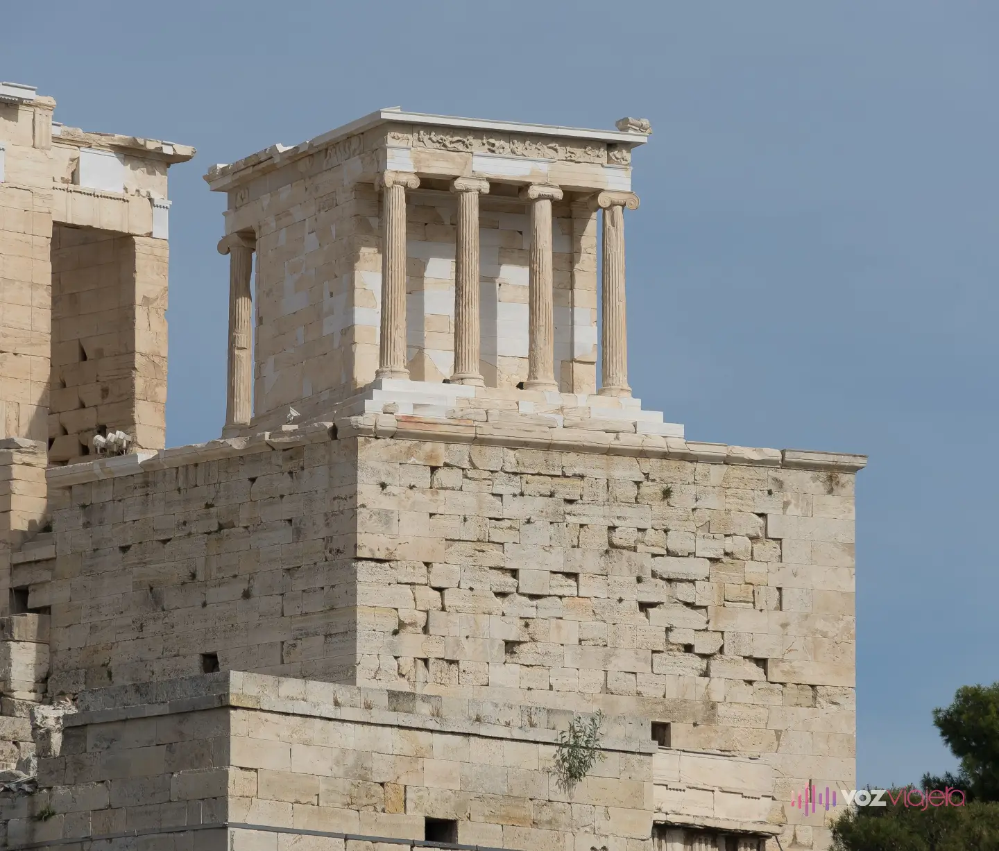 El Templo de Atenea Niké encaramado en lo alto de su bastión, visto desde abajo: el muro de sillares del bastión ocupa la mitad de la imagen y el templo, mucho más pequeño, asoma encima; a la izquierda, el muro de los Propileos