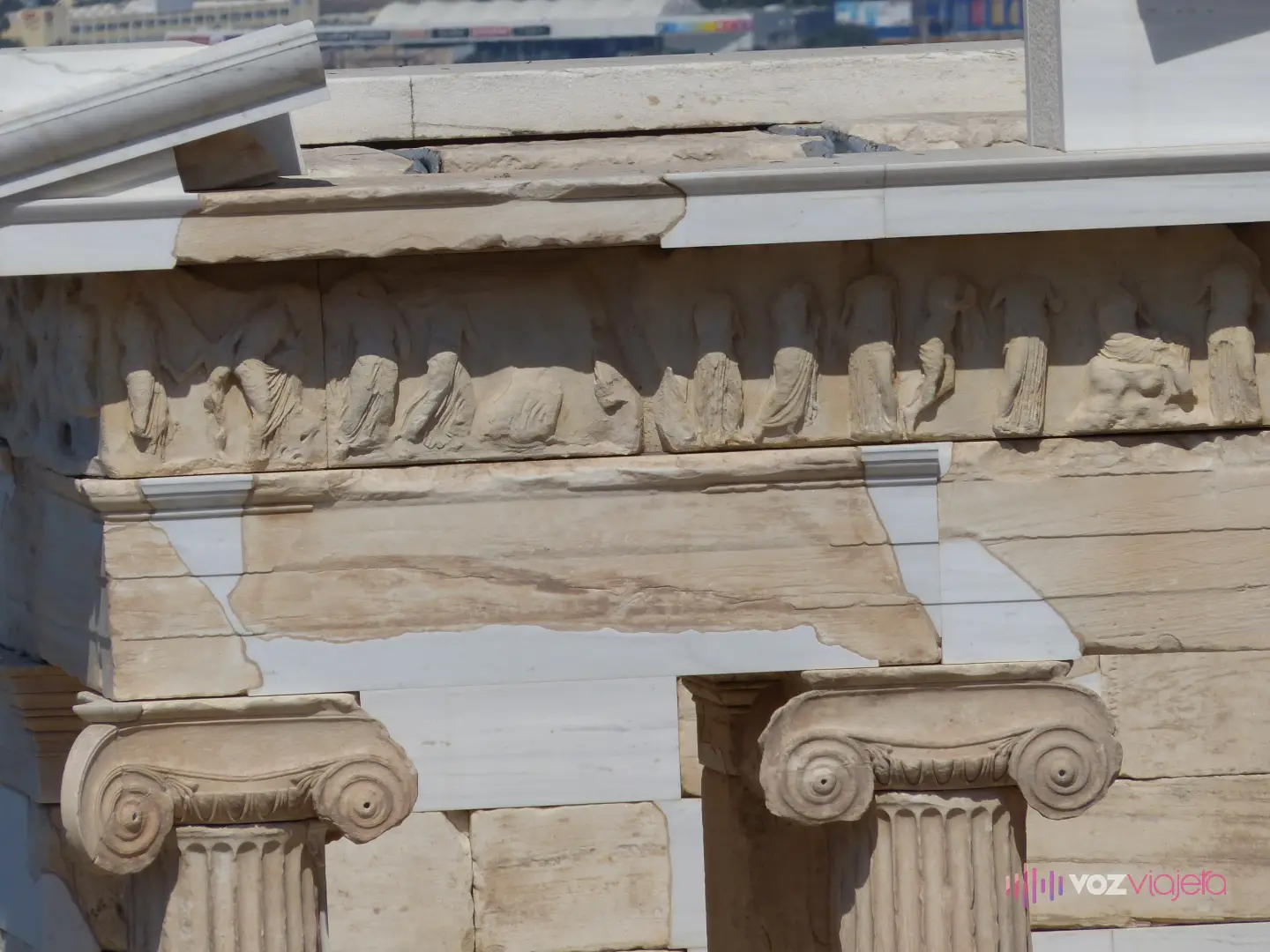 Detalle del friso del Templo de Atenea Niké visto de cerca: una banda estrecha de figuras vestidas con túnicas de pliegues muy marcados, una sentada y un caballo en el extremo derecho, sobre el arquitrabe y dos capiteles jónicos con sus volutas
