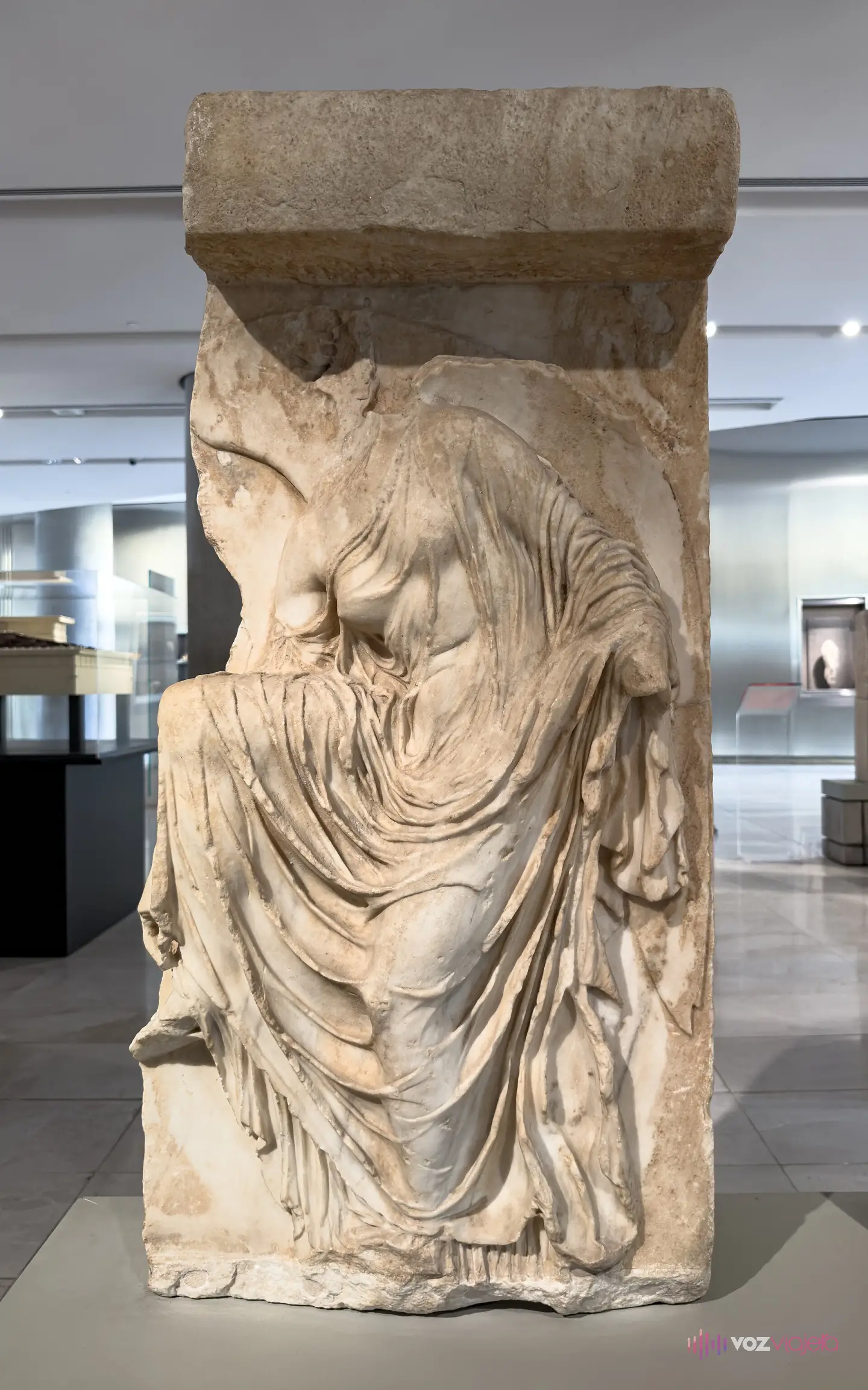 La placa de mármol de la Niké que se ata la sandalia, expuesta de pie en una sala del Museo de la Acrópolis: la figura se inclina hacia delante, le falta la cabeza y el vestido se le pega al cuerpo en pliegues finísimos que le resbalan del hombro