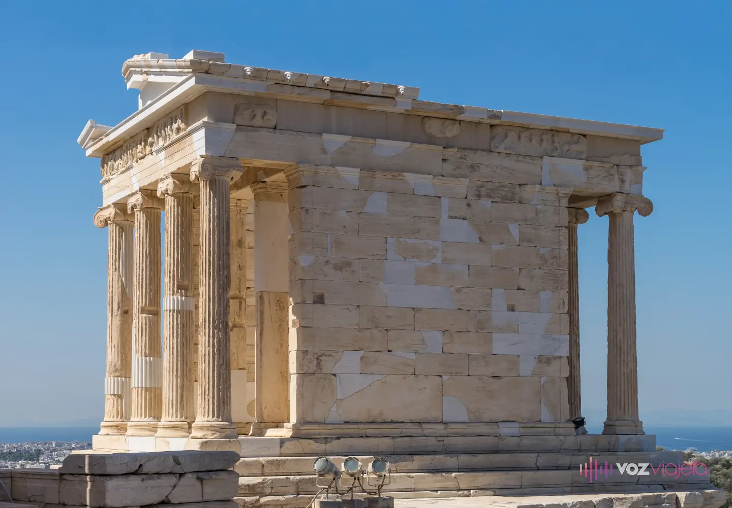 El Templo de Atenea Niké entero, visto desde una esquina: las 4 columnas jónicas de un extremo, el muro liso de la cela sin ninguna columna en el flanco y las del otro extremo asomando al fondo, con cielo despejado y el mar al horizonte