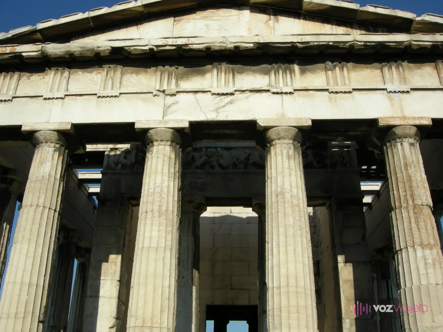 La fachada del Templo de Hefesto vista de frente y desde abajo, con el frontón triangular vacío, la hilera de triglifos y metopas del entablamento y las columnas dóricas acanaladas; entre ellas, en penumbra, asoma el friso esculpido