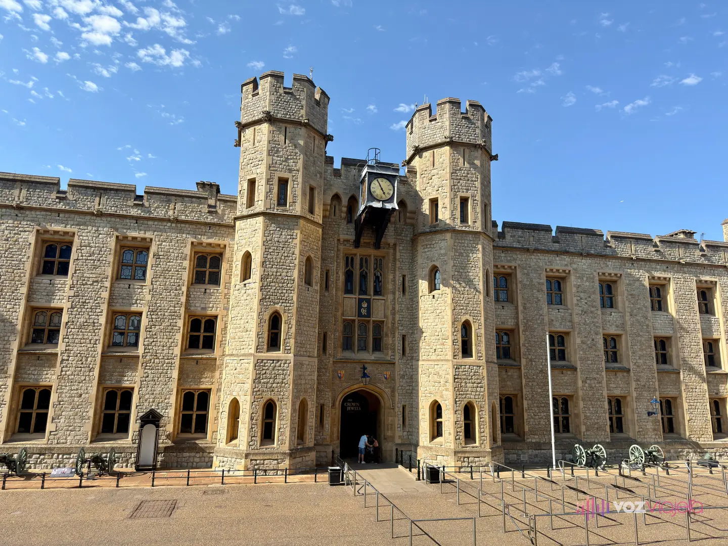 Fachada del cuartel de Waterloo en la Torre de Londres, de piedra clara y almenada, con 2 torres octogonales a los lados de la puerta; sobre el arco de entrada se lee «The Crown Jewels», encima cuelga un reloj negro y delante del edificio hay vallas metálicas en zigzag para la cola y cañones antiguos