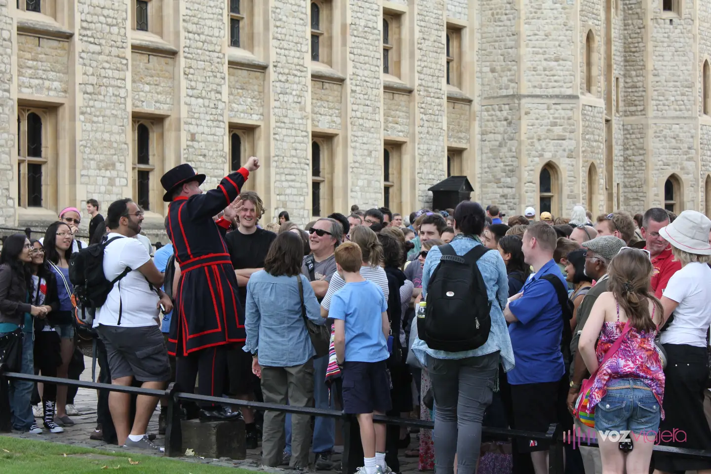 Un Yeoman Warder con el uniforme azul oscuro y rojo y sombrero Tudor, subido a un escalón, levanta el brazo mientras habla a un grupo numeroso de visitantes junto a un edificio de piedra de la Torre de Londres