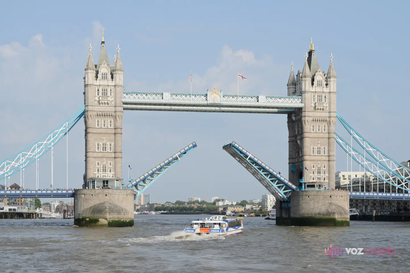 Tower Bridge abierto en un día soleado: las 2 básculas de la calzada levantadas en diagonal entre las torres y un barco blanco y azul cruzando por debajo sobre el Támesis