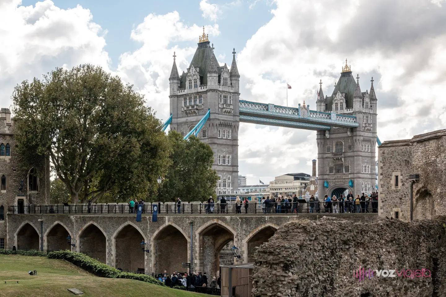 Las 2 torres de Tower Bridge asomando tras los muros de piedra y los arcos de la Torre de Londres, con árboles, césped y visitantes en el paseo de la muralla