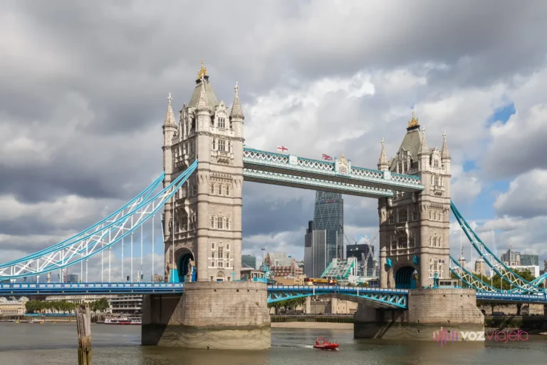 Tower Bridge entero con el Támesis en primer plano en un día de nubes: sus 2 torres de piedra unidas por las pasarelas altas, los tirantes pintados de azul claro y una lancha roja bajo el puente