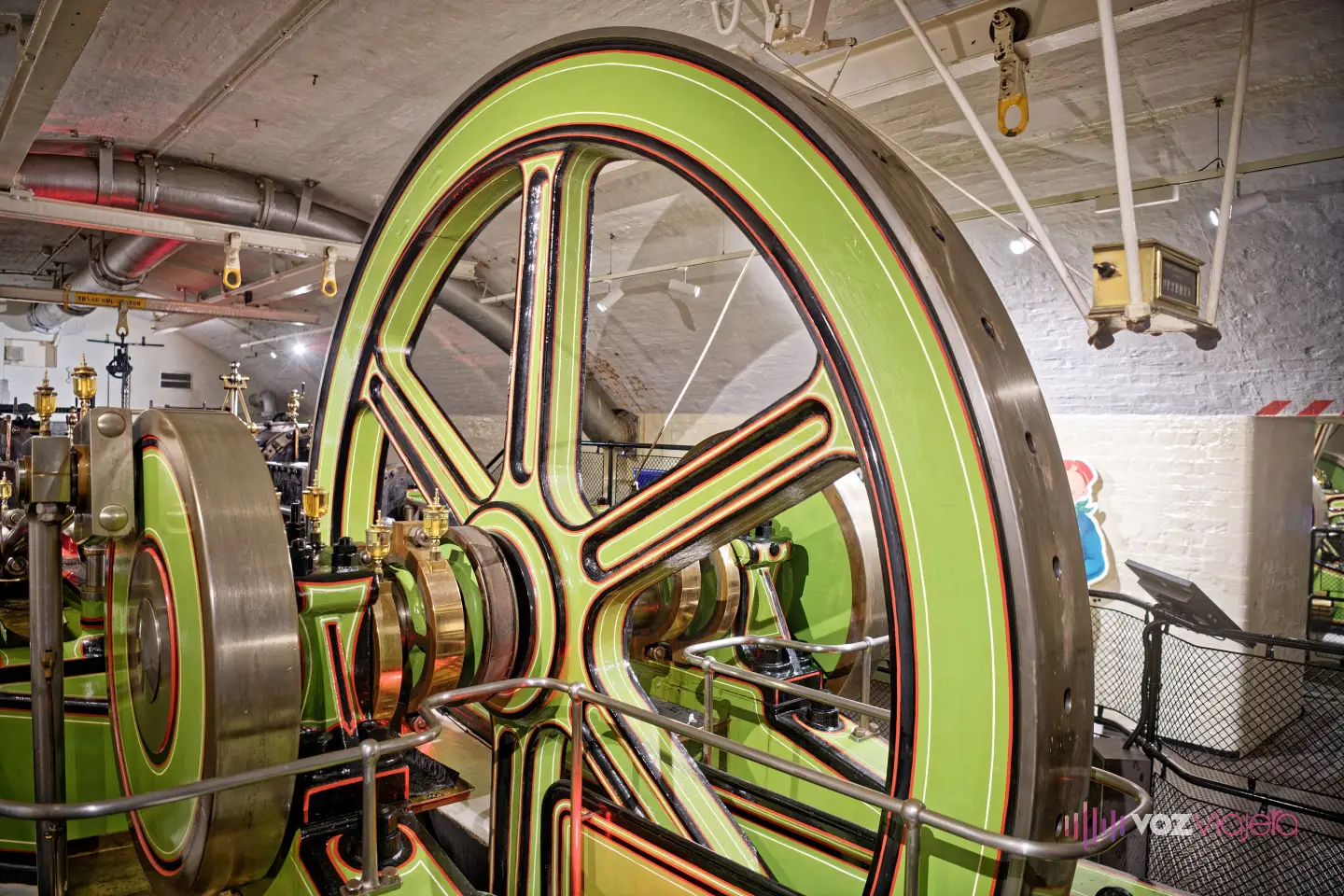 Gran volante de una máquina de vapor pintado de verde con filetes rojos y negros en la sala de máquinas victoriana de Tower Bridge, bajo un techo abovedado de ladrillo blanco con tuberías