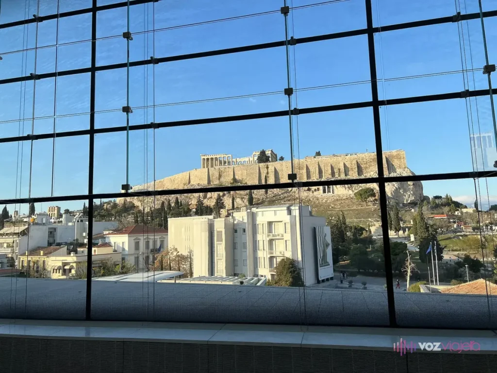 La roca de la Acrópolis con el Partenón en lo alto, vista a través de la retícula de una gran cristalera del Museo de la Acrópolis, sobre edificios blancos de la ciudad