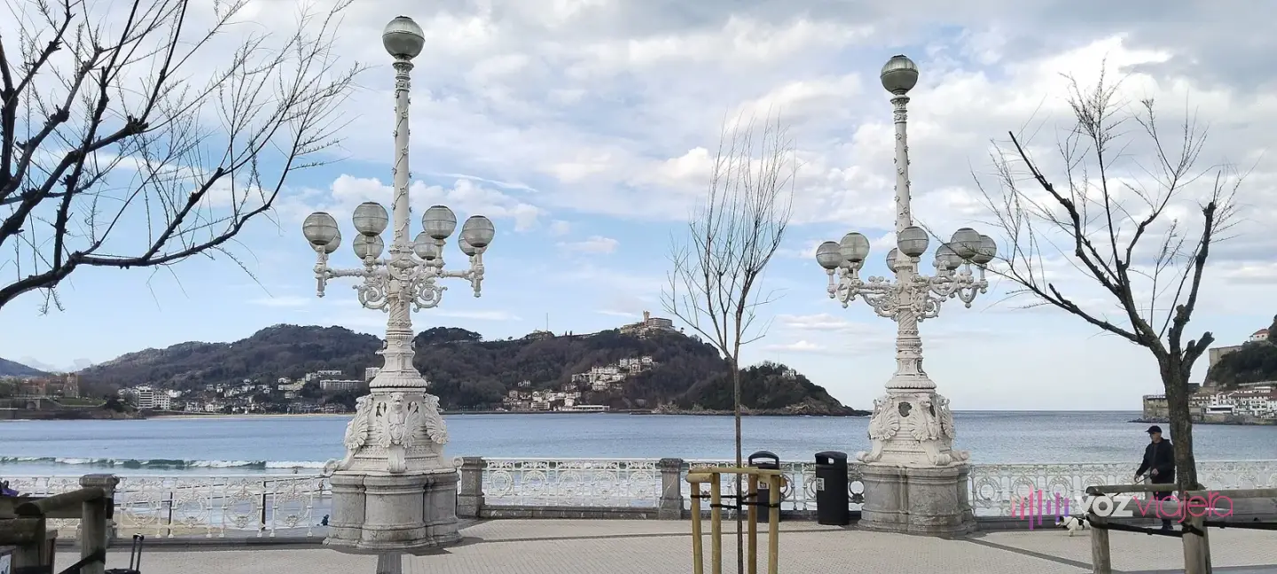 Dos farolas blancas de varios brazos y la barandilla blanca del paseo de La Concha, con la bahía y el monte Igeldo al fondo