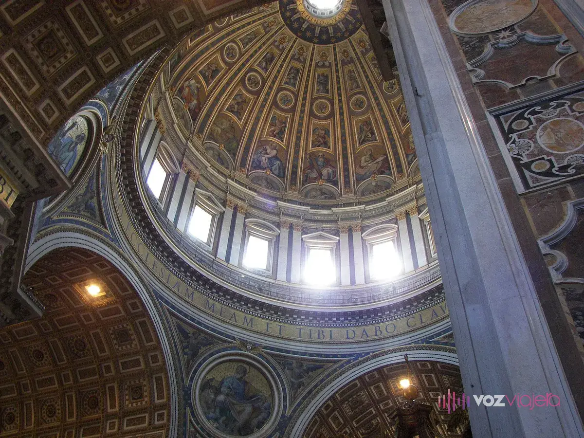 El interior de la cúpula de San Pedro visto desde abajo: el anillo de ventanas del tambor, la cúpula nervada cubierta de mosaicos sobre fondo dorado y, en su base, un friso con letras latinas grandes
