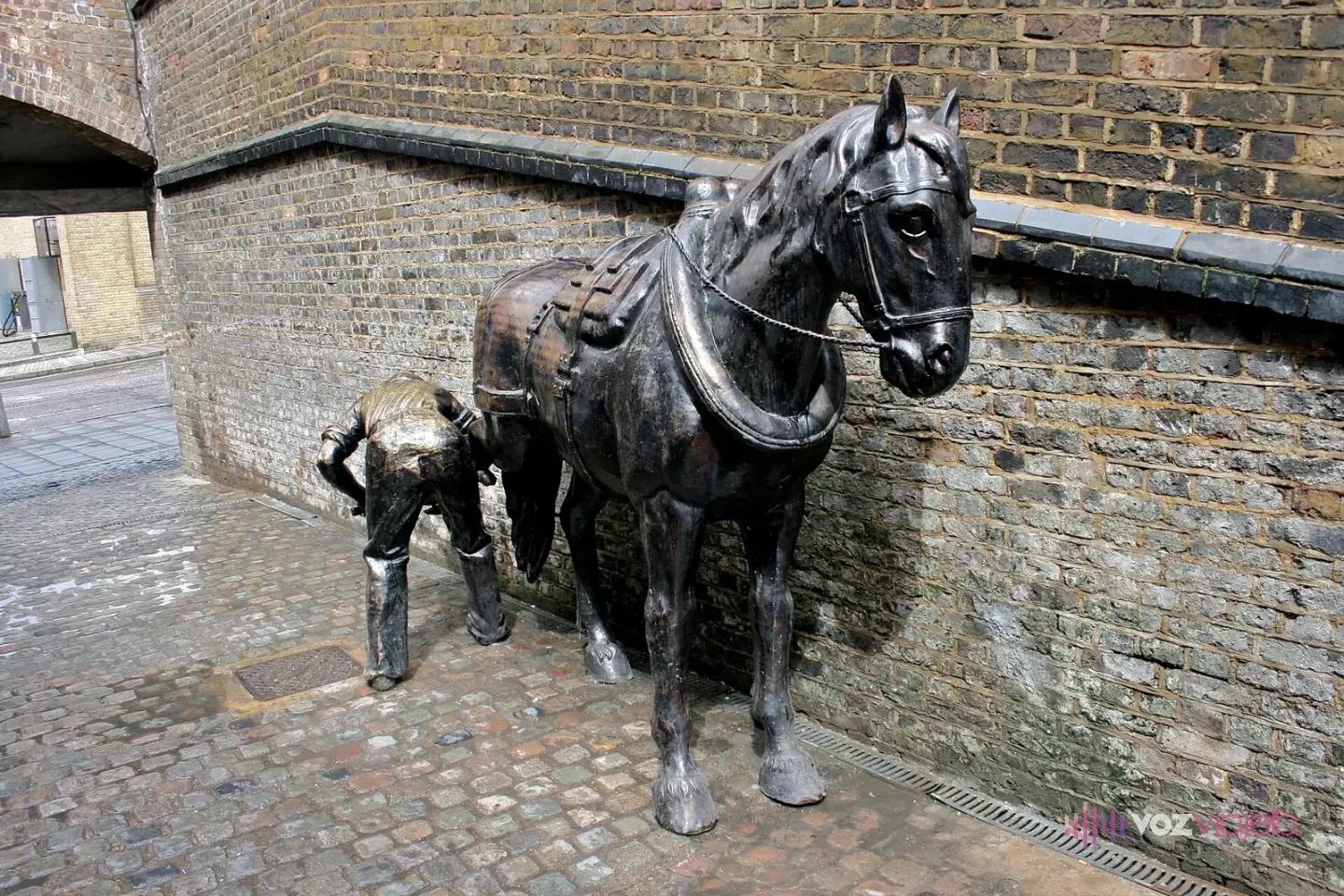 Escultura de bronce de un caballo de tiro con arnés y un herrero que le sujeta la pata, junto a un muro de ladrillo del Stables Market