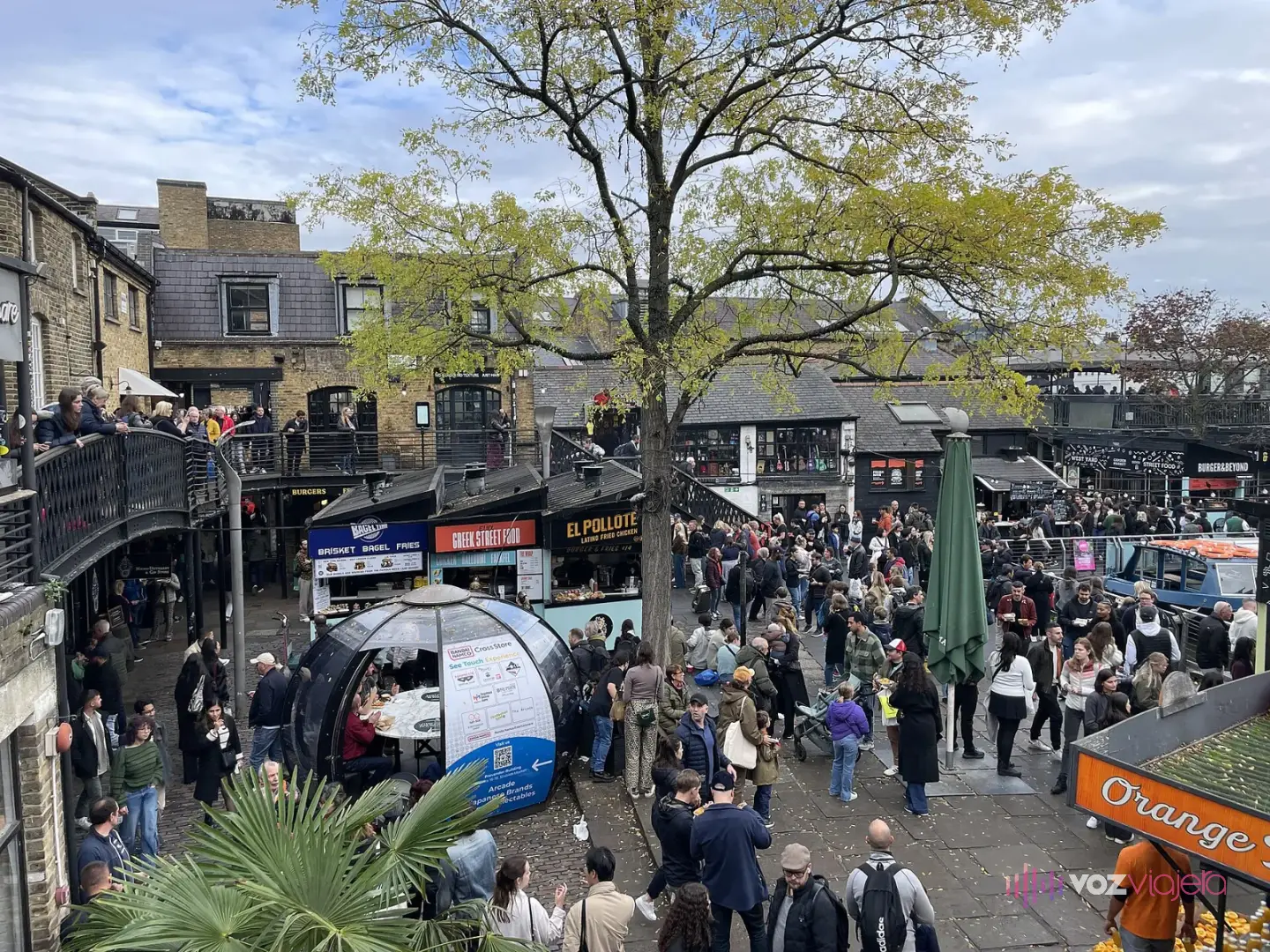 Patio de Camden Lock Market lleno de gente entre puestos de comida, con un árbol en el centro y los almacenes de ladrillo detrás
