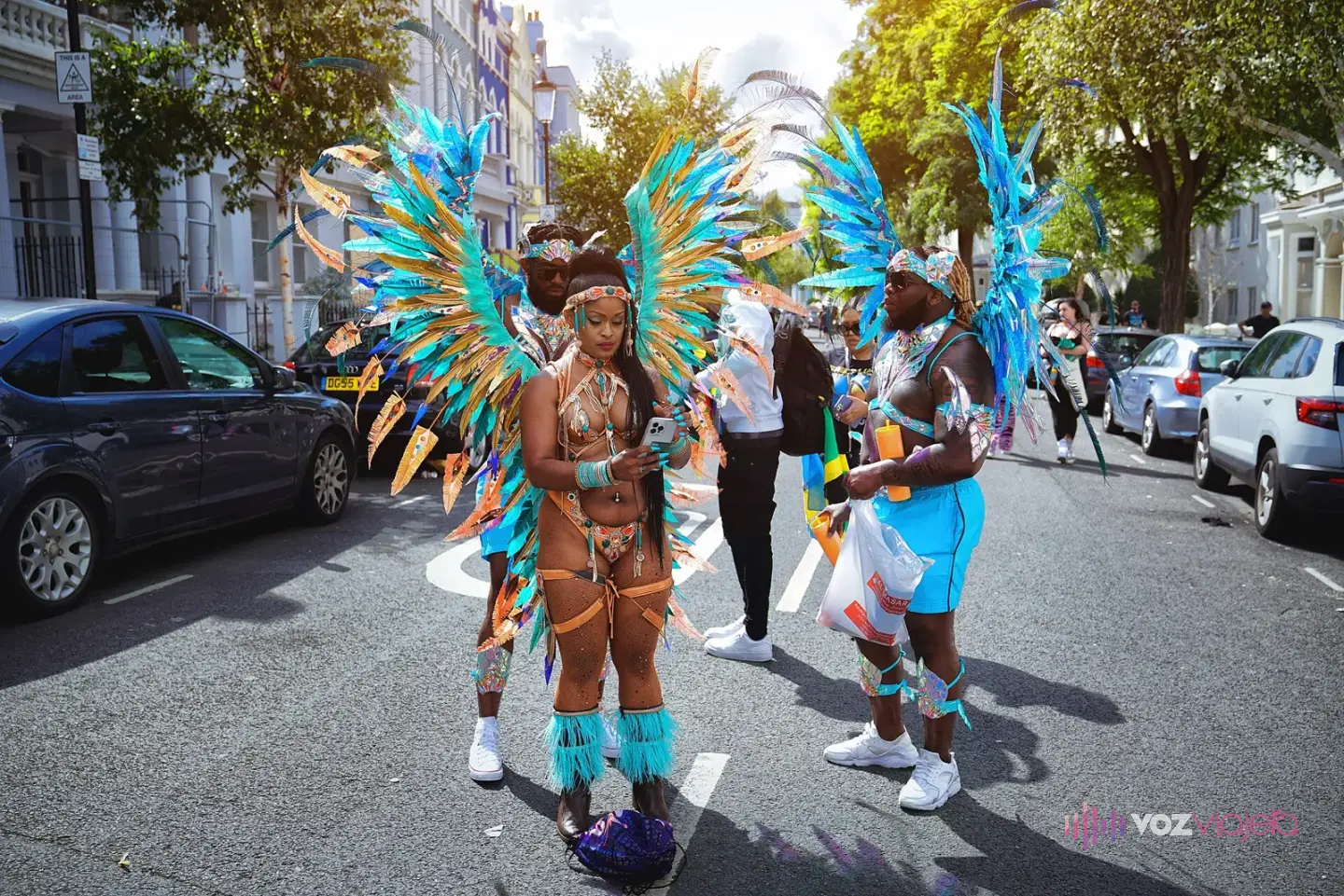 Tres integrantes de una comparsa del Carnaval de Notting Hill, una mujer y dos hombres, con alas de plumas turquesa y naranja, de pie en una calle con casas blancas y árboles