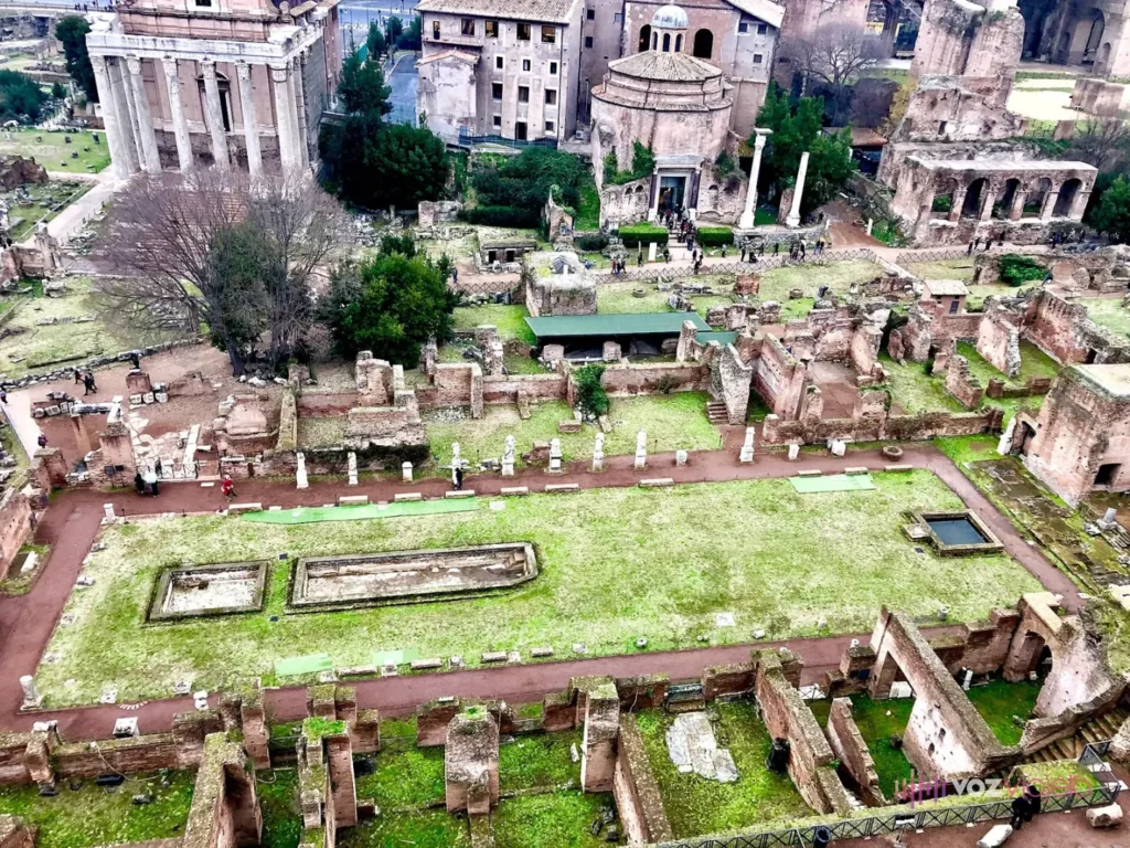La Casa de las Vestales vista desde el Palatino: un largo césped con estanques rectangulares entre muros de ladrillo y una fila de estatuas blancas sobre pedestales, con el templo redondo de Rómulo y las columnas del Templo de Antonino y Faustina detrás