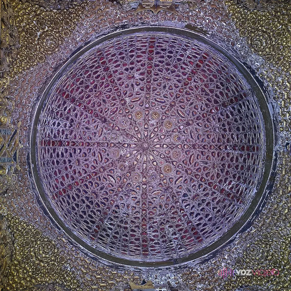 Cúpula de la escalera principal de la Casa de Pilatos vista desde abajo: media naranja de lacería en tonos morados y rojizos, rodeada de mocárabes dorados