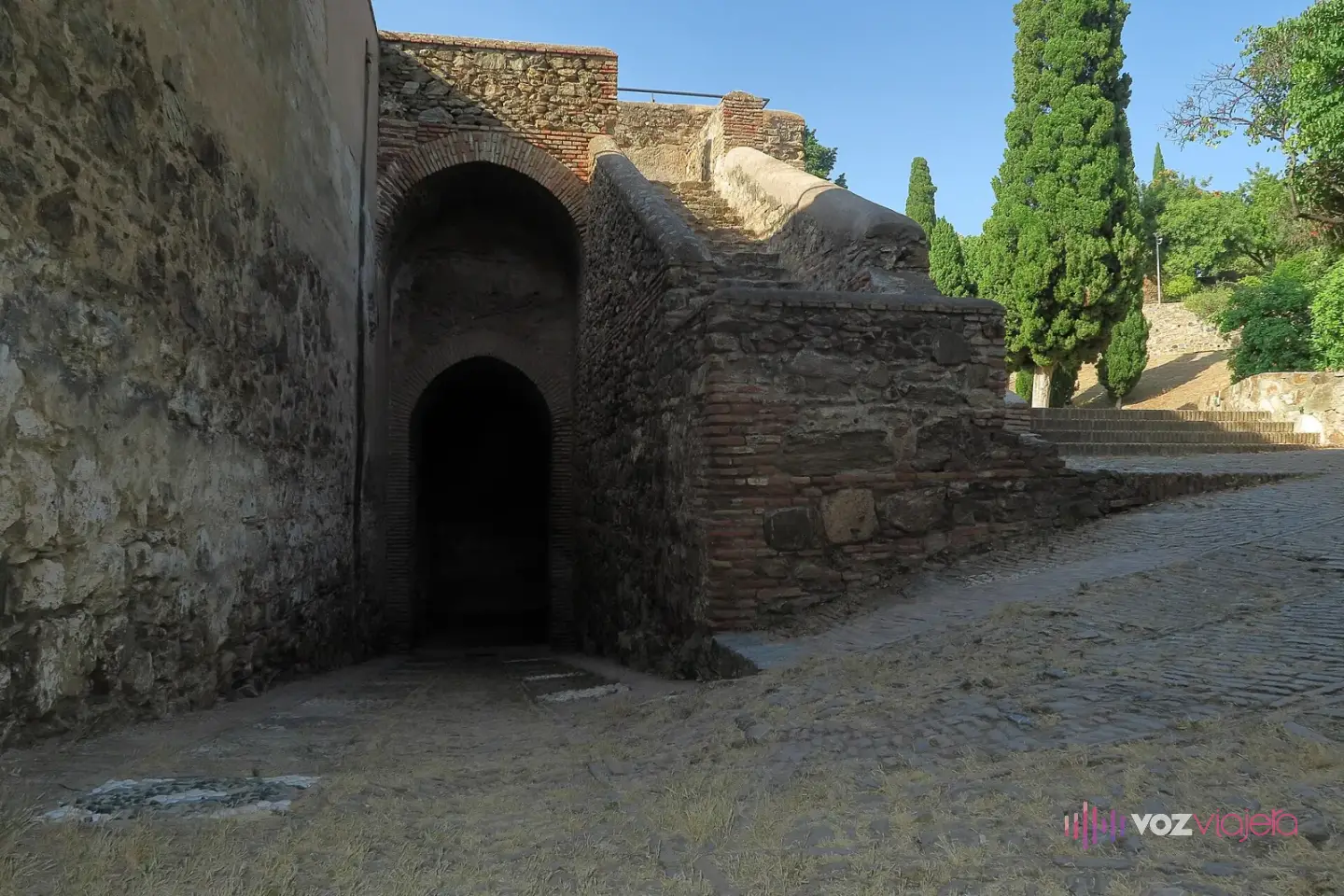 Puerta en arco abierta en un torreón de piedra y ladrillo del castillo de Gibralfaro, con una rampa empedrada y cipreses detrás
