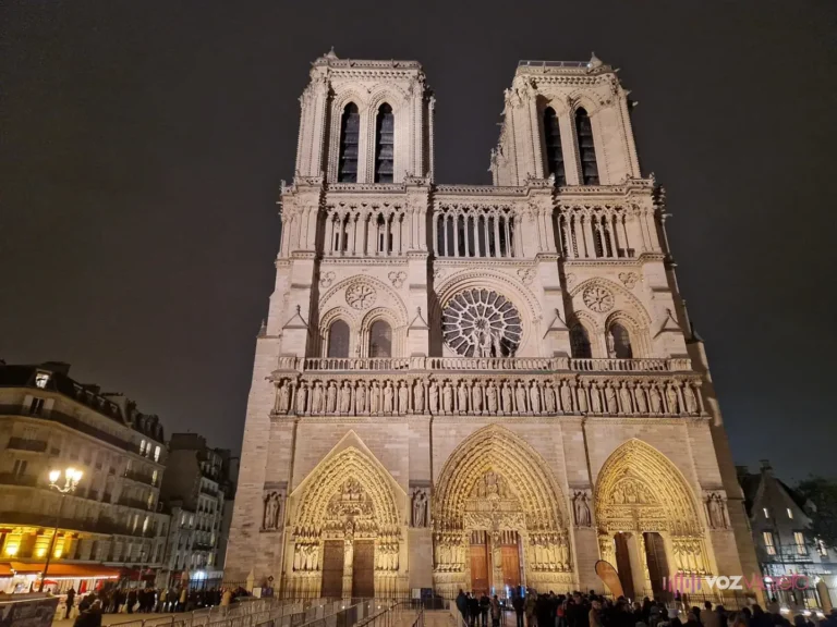 Fachada oeste de Notre Dame de París iluminada de noche, con sus dos torres, el rosetón central y los tres portales, y gente en la plaza