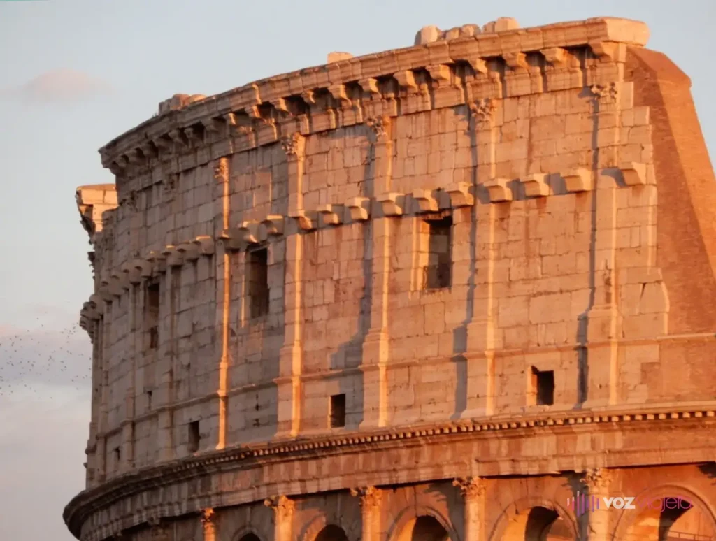 Lo alto del muro exterior del Coliseo al atardecer: el ático con sus ventanas cuadradas y ménsulas de piedra, sobre los arcos del tercer piso
