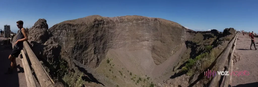 Panorámica del cráter del Vesubio desde el sendero: una gran hoya de paredes empinadas de roca parda y gris en capas, una barandilla de madera en el borde, un caminante apoyado a la izquierda y gente en el camino de grava a la derecha