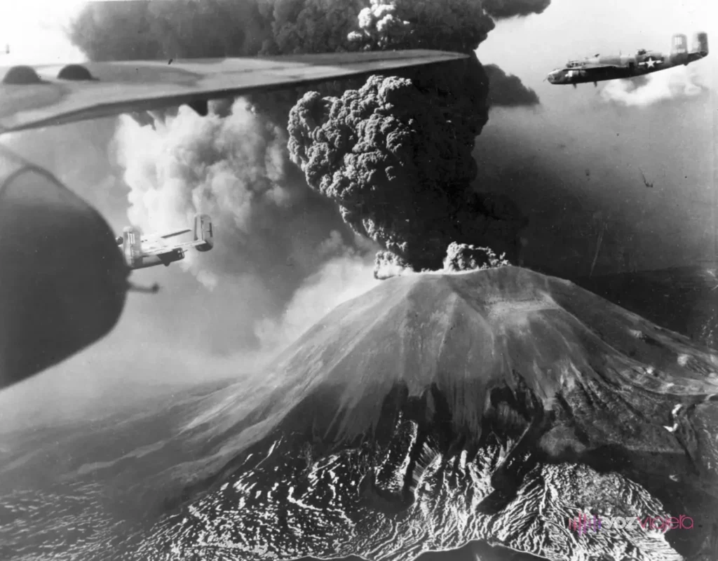 Foto aérea en blanco y negro del Vesubio en erupción en marzo de 1944: una columna oscura de ceniza sale de la cima del cono y pasan bombarderos B-25 estadounidenses, con el ala de otro avión en primer plano