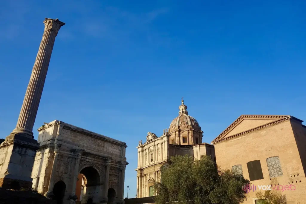 El extremo oeste del Foro Romano bajo un cielo azul: la alta Columna de Focas a la izquierda, el Arco de Septimio Severo detrás, la cúpula de la iglesia de los Santos Lucas y Martina y, a la derecha, la fachada sencilla de ladrillo de la Curia