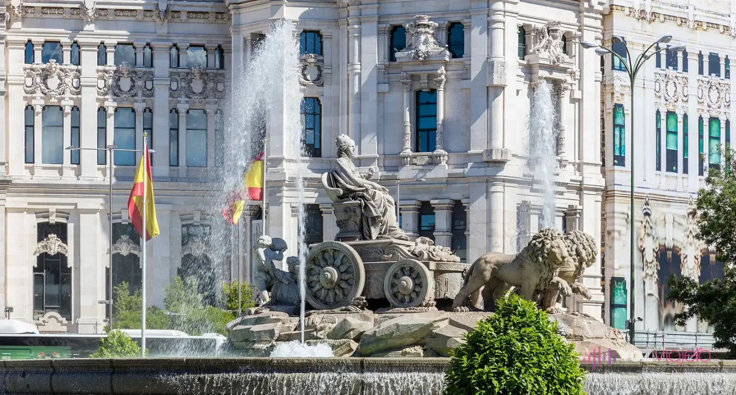 La Fuente de Cibeles con la diosa en su carro tirado por 2 leones y un surtidor alto, delante de la fachada blanca del Palacio de Cibeles con banderas de España