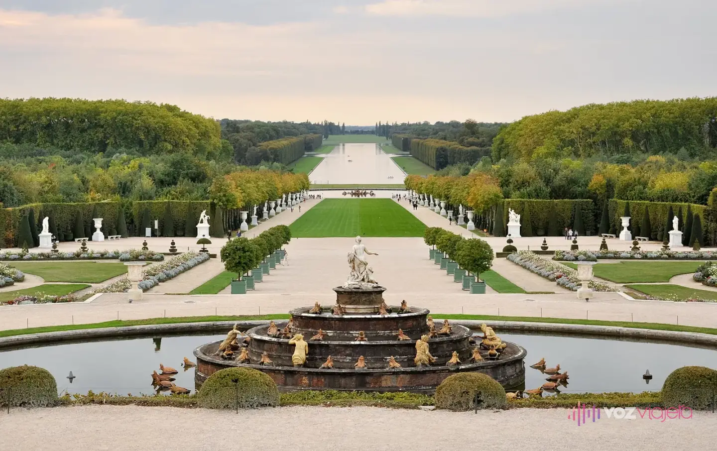 Los jardines de Versalles desde la terraza: una fuente con esculturas en primer plano, parterres y estatuas, y al fondo el Gran Canal entre arboledas