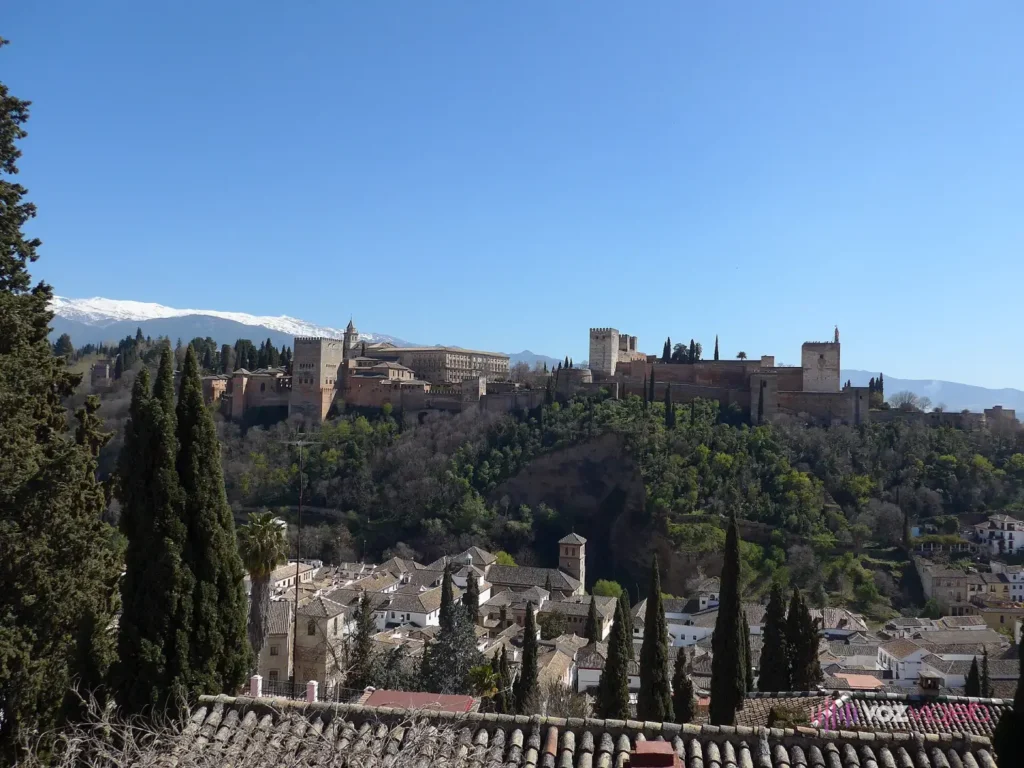 La Alhambra de Granada vista desde el Mirador de San Nicolás: murallas y torres rojizas sobre una colina arbolada, las casas blancas del Albaicín abajo y Sierra Nevada nevada al fondo